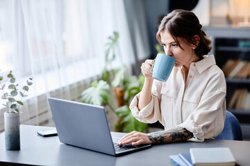 Portrait of tattooed young woman working from home and drinking coffee in morning, copy space