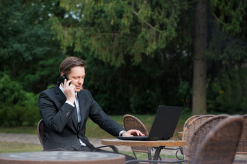 Busy working. Confident young caucasian businessman in formalwear working on laptop and talking on the mobile phone while sitting at the sidewalk cafe.