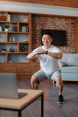 Domestic workout. Happy middle aged asian man doing deep squats during online training at home,...