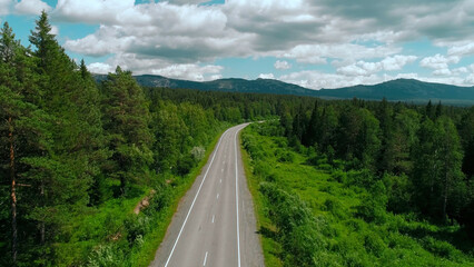 Obraz premium Aerial view of road across the forest on blue cloudy sky background. Scene. Straight long road bending along green trees and bushes.