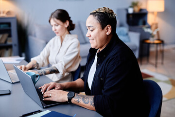 Side view portrait of two modern women using computers at home workplace together and smiling