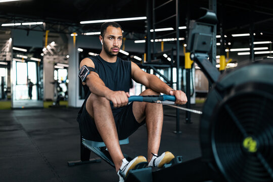 Sporty Young Black Man Exercising With Rowing Machine At Modern Gym