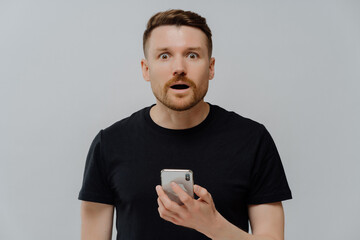 Horizontal shot of surprised startled bearded adult man holds mobile phone reacts on shocking news...