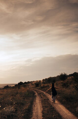 A girl walks along a deserted dusty road in a black T-shirt and a guitar in a case behind her back at sunset in summer
