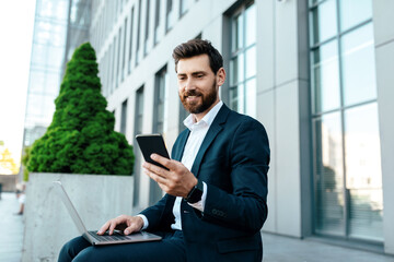 Happy young european businessman with beard in suit typing on phone and laptop, has chat with client