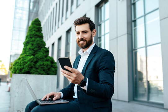 Happy young european businessman with beard in suit typing on phone and laptop, has chat with client