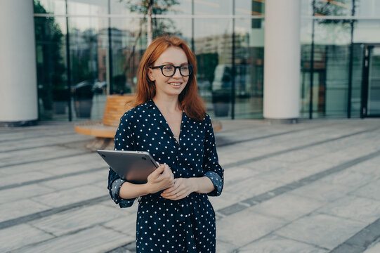 European female entrepreeur with red hair wears spectacles and polka dot dress holds modern laptop