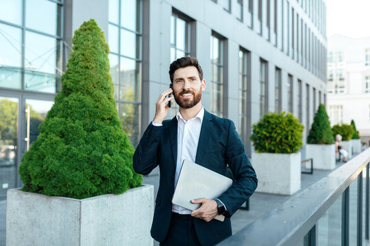 Busy smiling confident young caucasian businessman with beard in suit with laptop, call by smartphone
