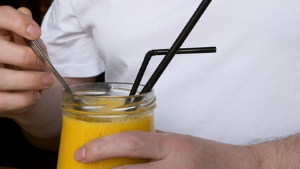 Close-up glass cup with fresh yellow mango smoothie, tasty pulp is eaten from it with a spoon....