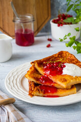 Finnish pancake pannukakku with redcurrant jam and sour cream or whipped cream on a white ceramic plate on a light wooden background. Scandinavian cuisine