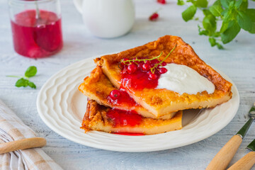 Finnish pancake pannukakku with redcurrant jam and sour cream or whipped cream on a white ceramic plate on a light wooden background. Scandinavian cuisine
