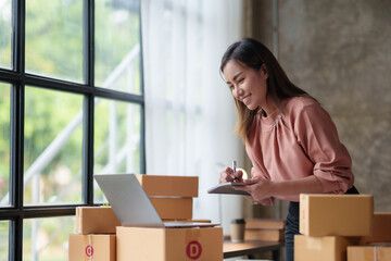 Smiling Asian online business owner prepares parcels and checks online order for delivery to customer on his laptop computer. Online selling concept.