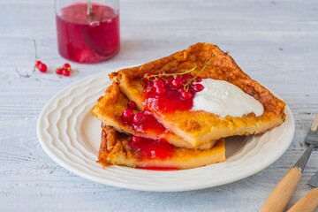 Finnish pancake pannukakku with redcurrant jam and sour cream or whipped cream on a white ceramic plate on a light wooden background. Scandinavian cuisine
