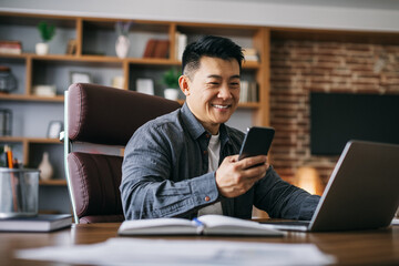 Happy handsome mature asian businessman typing on smartphone at workplace at laptop in home