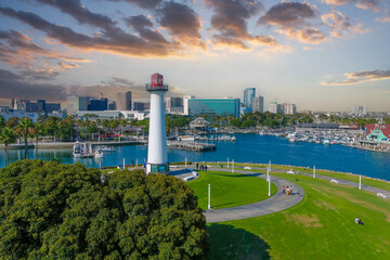 a gorgeous summer landscape in the park with a tall white and red lighthouse surrounded by lush green palm trees and grass with skyscrapers in the city skyline and blue ocean water at sunset