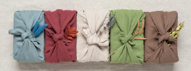 Gifts wrapped in colored fabric in a line on a beige textile linen background. Gifts in furoshiki style. Zero waste concept. Top view, selective focus. Banner
