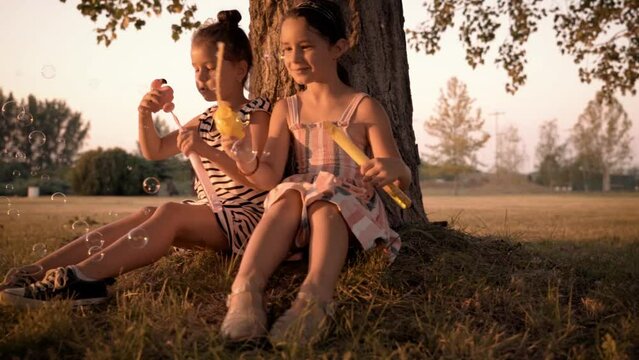 Two sweet little girls blowing a soap bubbles while sit leaned back on the tree. They have a fun with a soap bubbles and enjoying in a beautiful summer day.