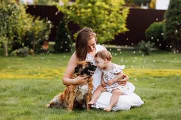 Fototapeta premium Family day, mother's day. Beautiful smiling young mom and baby daughter cuddling happy domestic dog on the backyard lawn.Mother with child girl are having fun with pet outdoors on summer holiday.