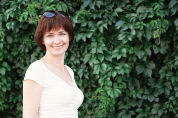 Woman with dark red hair stands and smiles in front of a wall covered in leaves. A woman lokking at the camera and smiling. Space for text