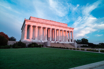 Pink light from sunrise illuminates the east-facing columns and exterior of the Lincoln Memorial in Washington, DC. Several sightseers sit on the marble steps. - obrazy, fototapety, plakaty