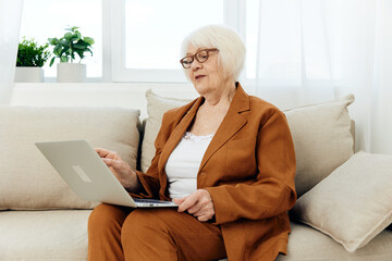 a joyful elderly woman is mastering new technologies using the Internet on a laptop while sitting on the couch at home