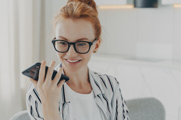 Beautiful positive red-haired woman in spectacles holding mobile phone and sending audio message