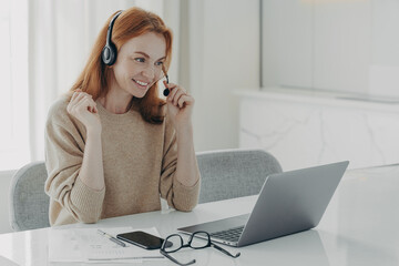Pleasant smiling redhead woman in headset communicating with colleagues during video call on laptop
