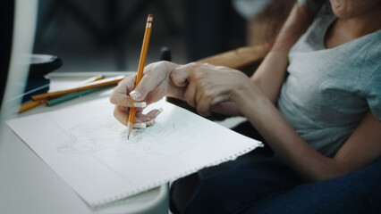 Woman with disability in a wheelchair sitting at the desk and drawing a sketch, with pencil on a sheet of paper and records it on video or makes a live stream on the social network
