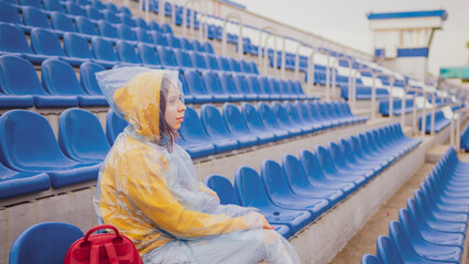 Young woman in raincoat sitting on stadium bleachers alone in rainy weather. Female spectator in...