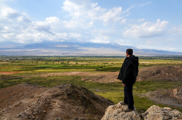 Naklejka na ściany i meble Rear view of a man standing, looking and enjoying of a scenic landscape a valley with mount Ararat hidden by clouds