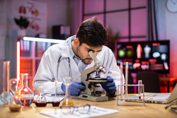 Young indian male researcher looking at the microscope analyze science, forensics, microbiology, biochemistry, genetics, oncology, laboratory.