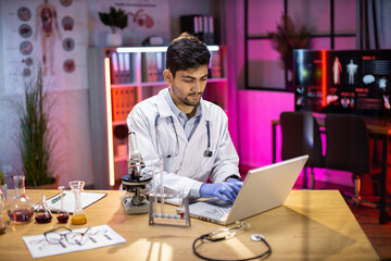 Medical research laboratory: portrait of a attractive male scientist using digital laptop computer, analysing liquid biochemicals in a laboratory flask. Advanced scientific biotechnology laboratory.