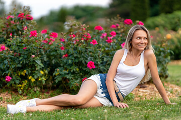 Naklejka na ściany i meble A beautiful, sensual and sexy blonde young lady in a white top and denim shorts posing near blooming roses in a garden
