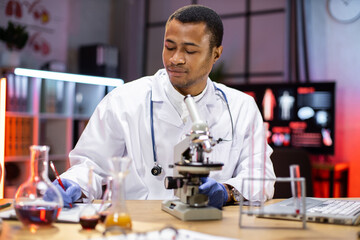 Fototapeta premium Professional african american man microbiologist or medical worker use test tube filling with blood and microscope writing research results in the laboratory.
