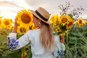 Portrait of young woman walking in blooming sunflower field at sunset smelling picking flowers....