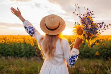 Back view of happy woman enjoying view in blooming sunflower field at sunset with bouquet of...