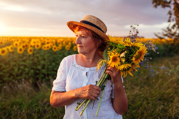 Senior woman holds bouquet of yellow sunflowers in summer field at sunset. Middle-aged woman...