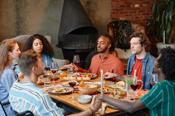 Diverse group of young people holding hands at table and saying grace during dinner party in cozy setting