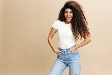 Good offer. Happy stylish curly Latin female in white t-shirt, smiling at camera, hold hands on pocket, say Yeah, isolated beige background. Copy space clothing fashion brands, free place for your ad