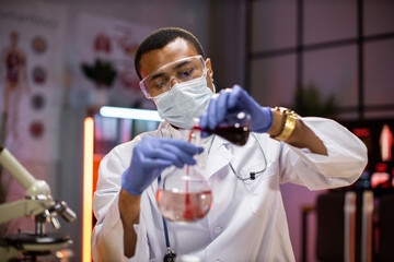 Science, chemistry, technology, biology and people concept - young african american male in protective mask and goggles scientist with test tube and microscope making research in clinical laboratory.