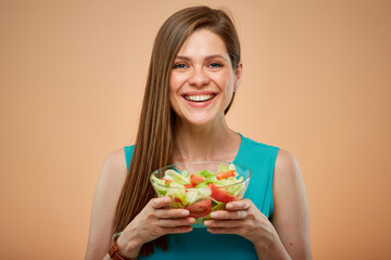 Young lady in azure dress holding green salad in glass bowl, isolated portrait on peach color background