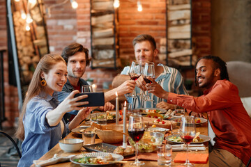Group of young friends taking selfie photo at table during dinner party and clinking glasses