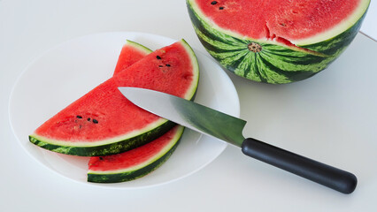 Close-up slices of fresh ripe red watermelon on a plate with a knife on the table on a white background, half of a watermelon. Ripe juicy summer berries, melon season. Macrophoto