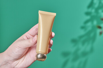Female hand holds cosmetic product in tube, glass bottle, lotion, anti-aging moisturizer, facial essential oil or serum on green background with light shadow nature. Beauty cosmetic skincare concept.