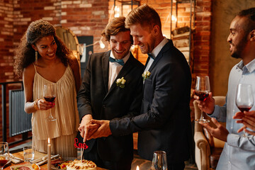 Portrait of young gay couple cutting cake together during wedding reception, same sex marriage