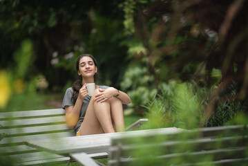 Cheerful young woman relaxing and breathing fresh air while sitting in balcony with nature view and drinking morning coffee and beverage