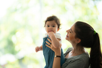 Cute little baby toddler playing and enjoying while in the arms of young mother hugging at home