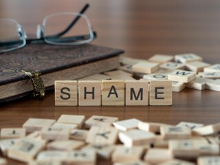 shame word or concept represented by wooden letter tiles on a wooden table with glasses and a book