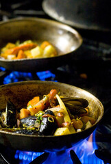 Root vegetables and seafood including mussles being cooked in a pan over an open flame on a stovetop.  Image features intentional blur due to shallow depth of field.