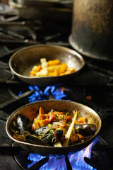 Root vegetables and seafood including mussels being cooked in a pan over an open flame on a stovetop.  Image features intentional blur due to shallow depth of field.